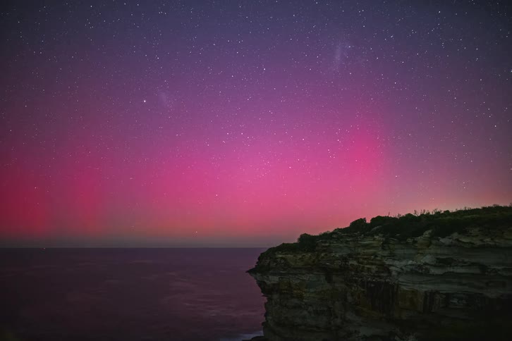 Time-lapse of a magnificent magenta aurora glowing over a calm sea and a rocky cliff under a starry night sky
