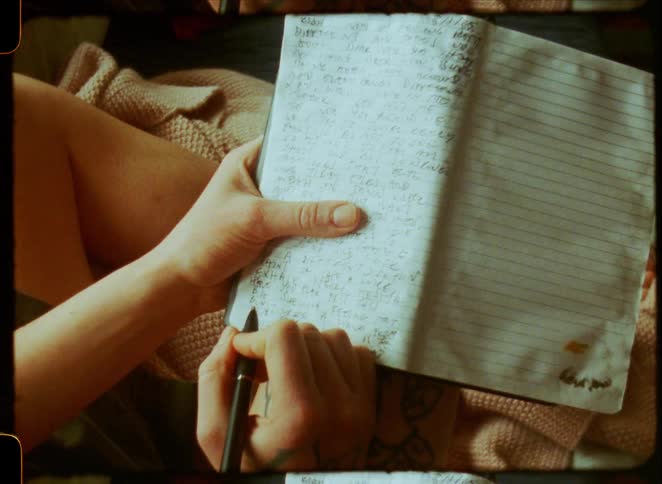 An introspective moment captured as a person with tattoos writes their thoughts in a journal with warm sunlight moving across the page