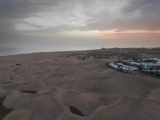 An aerial time-lapse of vast sand dunes along a coastline with a resort town under a cloudy sunset sky