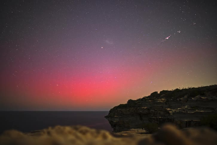 A celestial timelapse shows the beautiful Aurora Australis glowing in pink and purple over the ocean from a cliffside