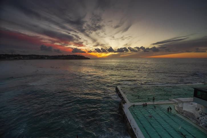 A stunning time-lapse of a vibrant sunrise over Bondi Beach, showing people swimming in the famous Icebergs Pool as waves crash and surfers catch the first light