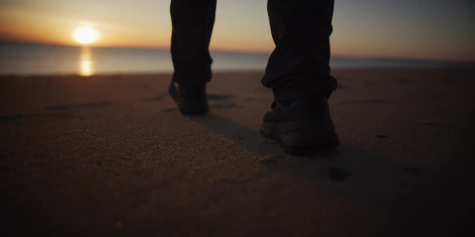 Close up slow motion of feet in sneakers walking on the sand towards the ocean during a golden sunset