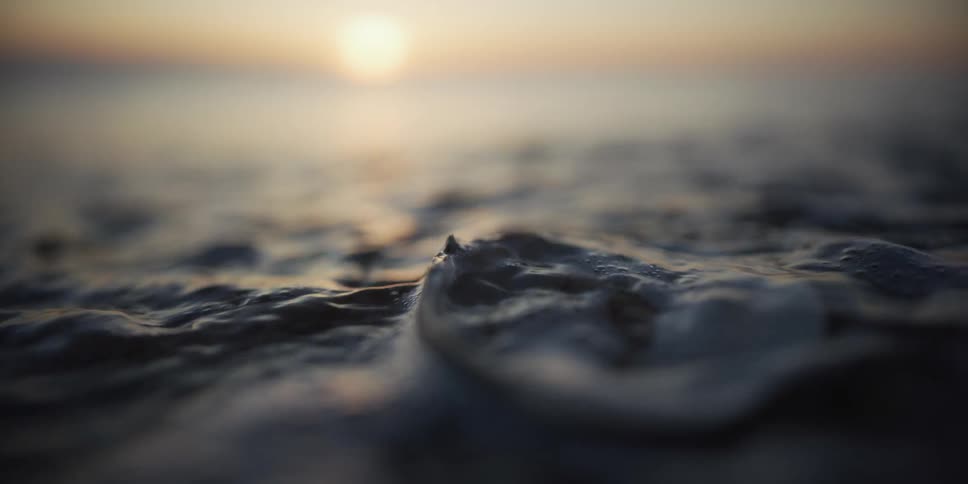 Serene macro footage shows a gentle tide washing over a smooth rock on a sandy beach at sunset