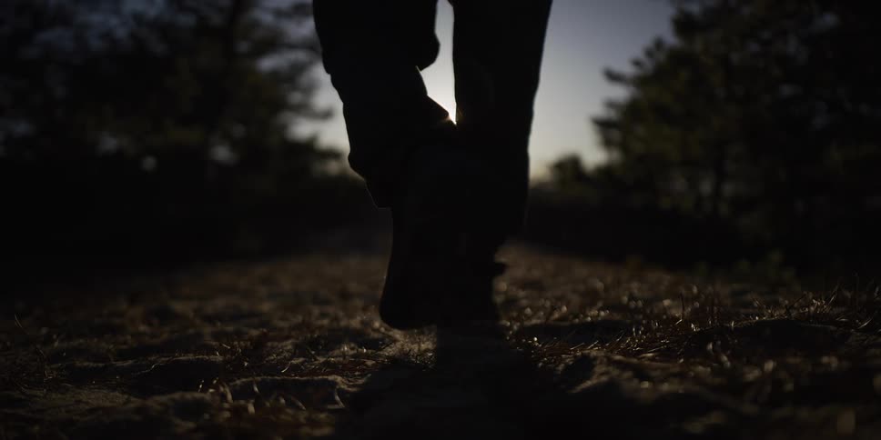 A low-angle slow-motion shot of a person's silhouette walking away from the camera towards the setting sun on a sandy trail