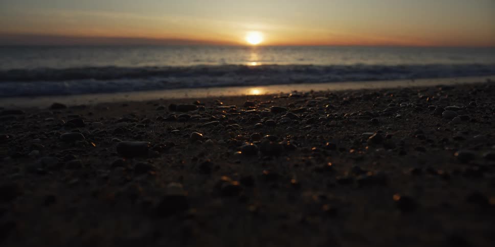 A serene low-angle close-up of a pebbly beach as gentle waves roll ashore during a beautiful golden sunset over the ocean