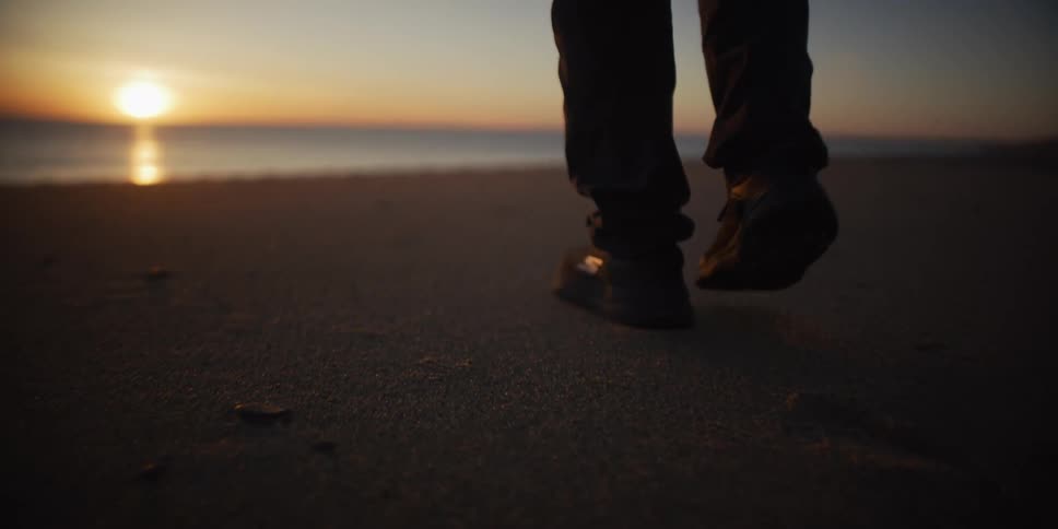 A person walks in slow motion on a sandy beach towards the ocean during a beautiful golden sunset