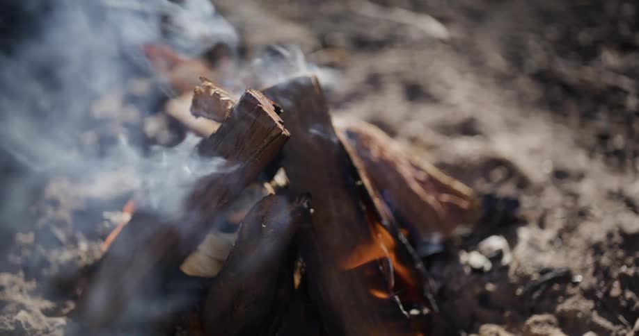A close-up slow-motion shot captures thick white smoke rising from a small smoldering campfire in the dirt