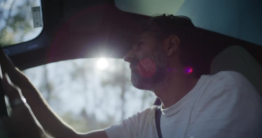 A relaxed middle-aged man with a beard drives his car as the bright sun creates a beautiful lens flare through the window