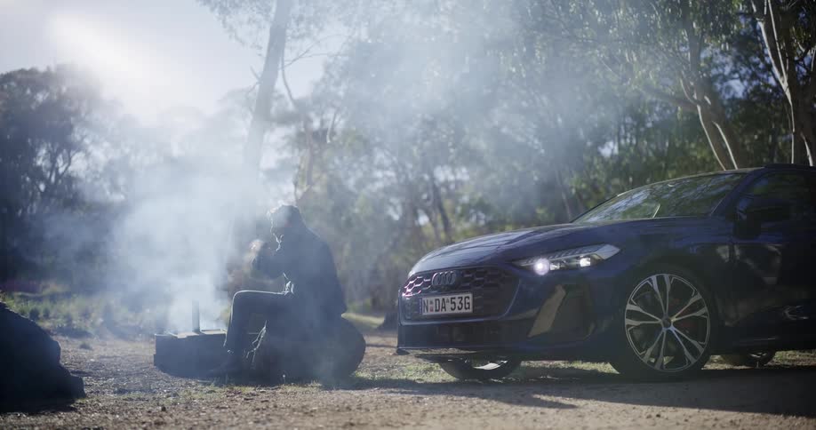 A man takes a break from his drive, sitting on a log and enjoying a drink by a smoky campfire in a sunlit forest next to his car