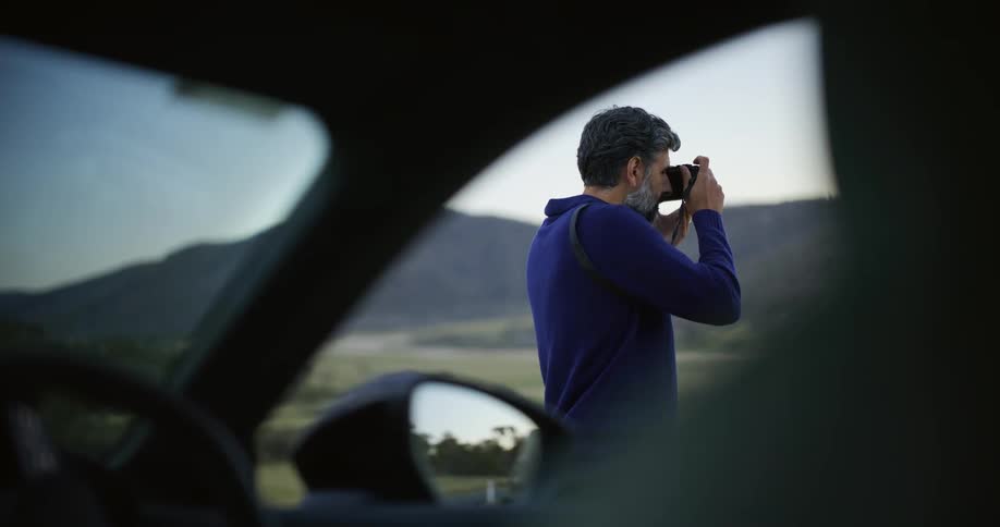 A thoughtful photographer, seen from inside a car, takes pictures of the rolling hills and mountains at sunset