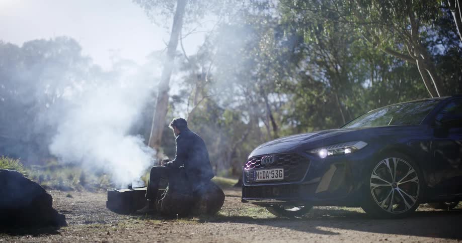 A man enjoys a peaceful morning, warming himself with a drink by a smoky campfire next to his car in the sunlit woods
