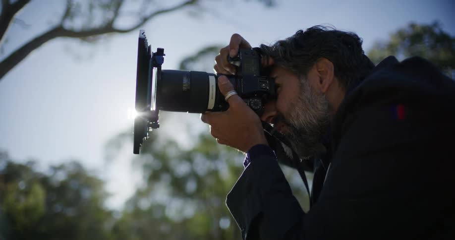 Close-up side profile of a professional photographer lining up a shot on his camera, with a bright sun flare illuminating the lens