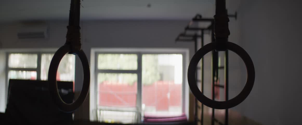 Still shot of wooden gymnastic rings hanging in a dimly lit, empty gym