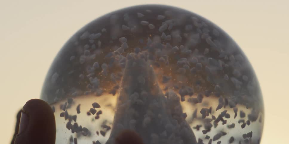 A hand holds up a snow globe with a mountain inside as flakes swirl in slow motion against a warm, hazy sunset