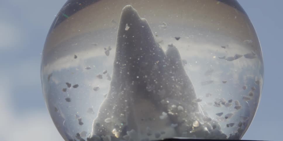 Tiny shells and glitter swirl like snow in slow motion around a miniature mountain inside a sunlit glass globe