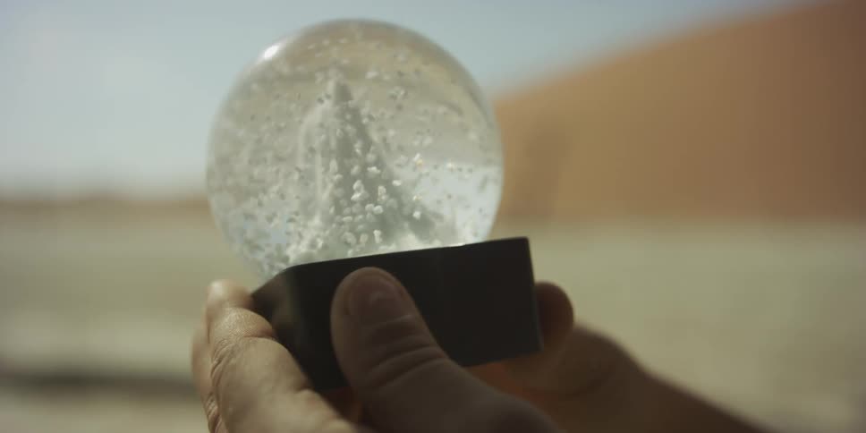 A close-up slow-motion shot shows a person shaking a snow globe with a miniature sand dune inside, set against a real desert landscape