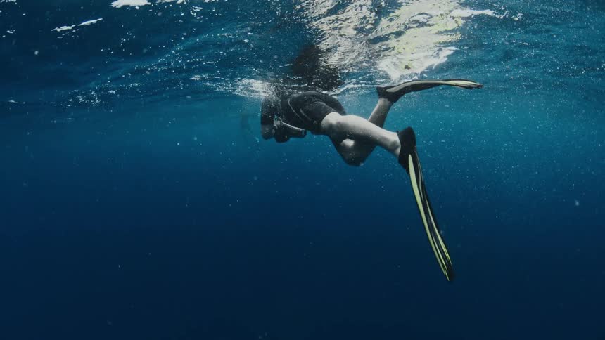 An underwater photographer in a wetsuit and fins gracefully glides through the deep blue ocean in slow motion