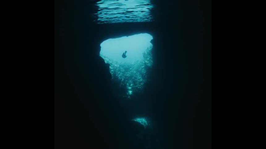 A lone diver explores a dark underwater cave, silhouetted by the light filtering through an opening from above