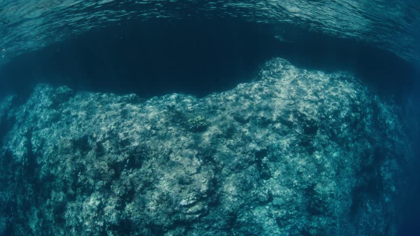A serene underwater view shows sunlight dancing across a large rock formation below the ocean's surface