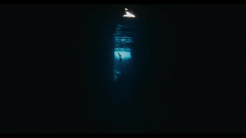 A silhouetted scuba diver floats on the surface inside a dark, mysterious underwater cave, illuminated by a single beam of light from above