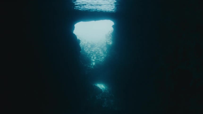 A serene underwater view from inside a dark cave, looking up at the sunlight filtering through an opening in the rock