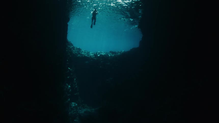 Looking up from the dark depths of an underwater cave at a lone snorkeler silhouetted on the ocean's surface