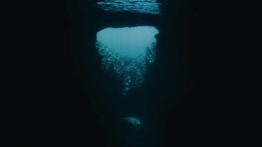 View from inside a dark underwater cave looking up towards the light at the surface as a free diver swims by