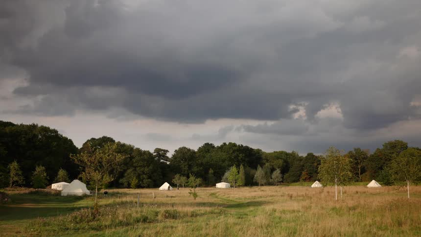A wide shot of a peaceful yurt campsite in a grassy field with a forest in the background, under dramatic dark storm clouds