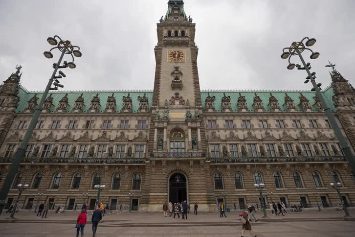 A slow push-in, time-lapse shot of the historic Hamburg City Hall as people walk through the square on a cloudy day