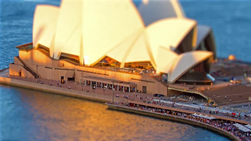 A slow zoom-out with a tilt-shift effect captures the iconic Sydney Opera House at sunset, with crowds of people enjoying the golden hour
