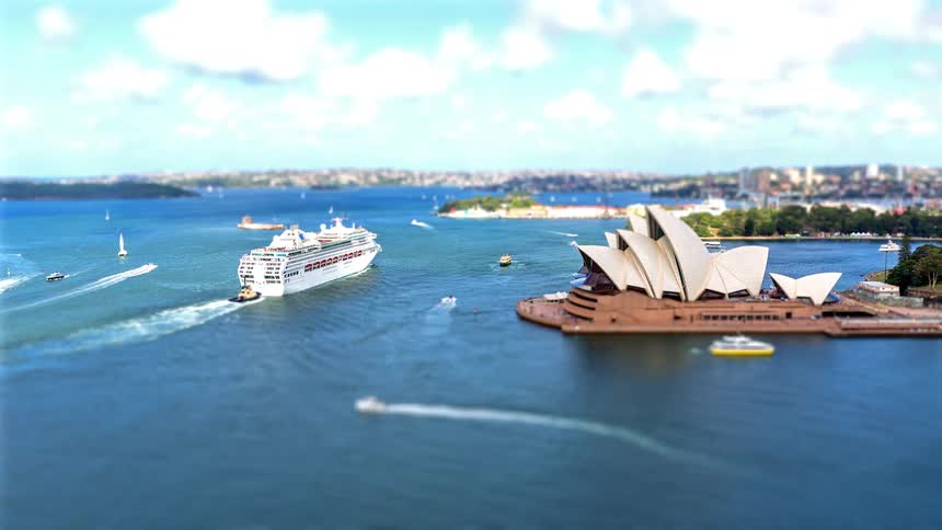 A tilt-shift view of a large cruise ship sailing past the iconic Sydney Opera House in a busy harbour on a sunny day