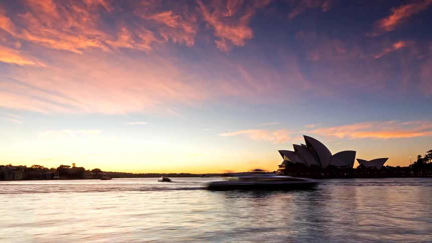 Time-lapse of a spectacular sunrise over the iconic Sydney Opera House, showing colorful clouds and bustling harbor traffic from night to day