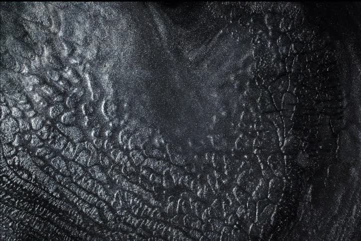 An abstract, dark metallic liquid with sparkling glitter slowly flows and settles into a textured pattern