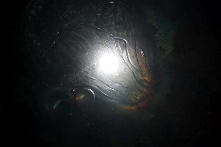 A bright orb of light shines through a slowly morphing dark, oily liquid creating an otherworldly effect