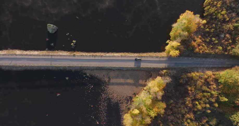 Top-down aerial footage of a car driving on a road built over a misty lake, flanked by vibrant autumn forests