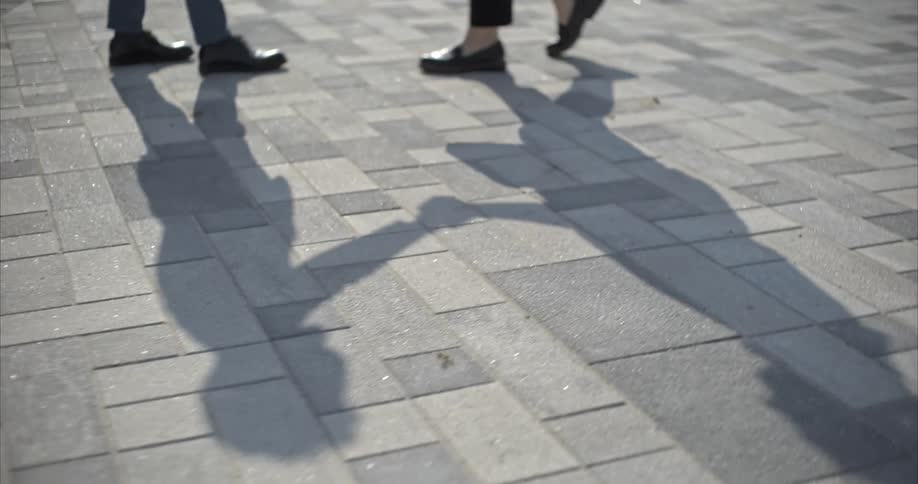 A slow-motion close-up captures the shadow of a man and woman holding hands on a paved sidewalk