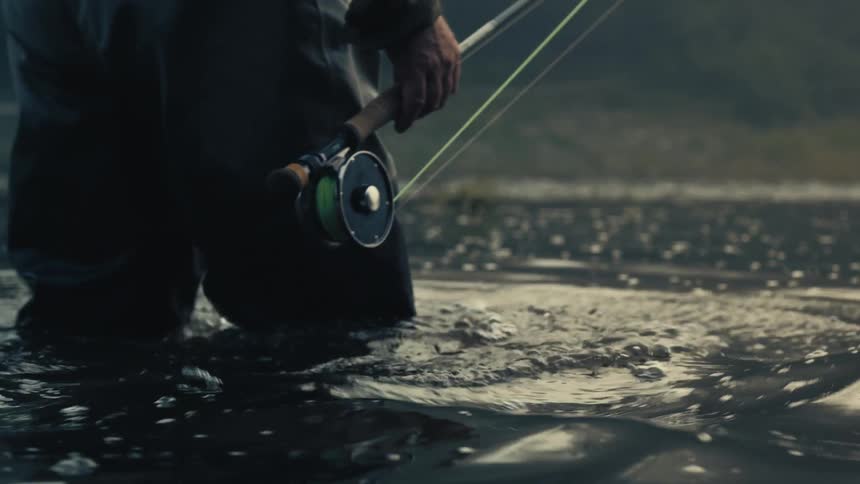 Close-up slow-motion shot of a fly fisherman in waders walking through a calm river