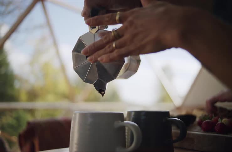 A person pours fresh coffee from a silver Moka pot into a mug during a rustic glamping breakfast in slow motion