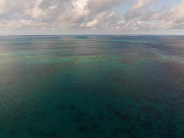 Vast aerial shot pulls back to reveal an expansive turquoise ocean with a beautiful coral reef system under a cloudy sky