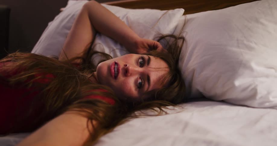 A dazed young woman with messy hair lies in bed and stares blankly into the camera in a vulnerable selfie-style shot