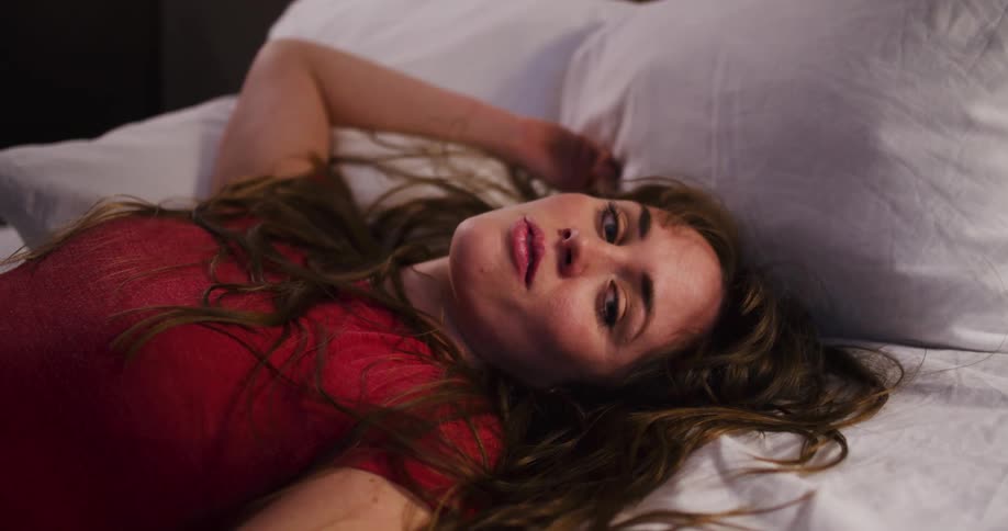 A woman with a fearful expression lies in bed staring into space as the camera slowly pulls away