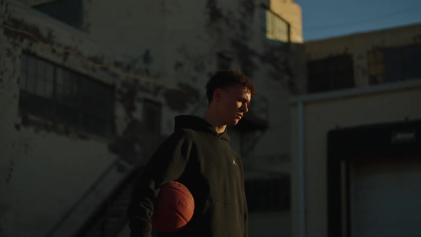 A determined young man in a black hoodie holds a basketball while contemplating in a shadowy urban alley at sunset