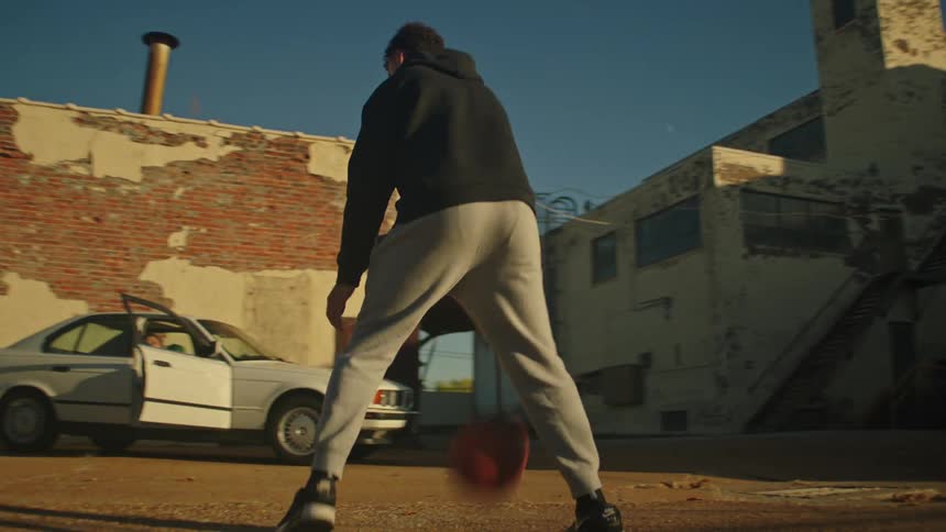 Cinematic footage of a young man dribbling a basketball in a run-down urban lot at golden hour