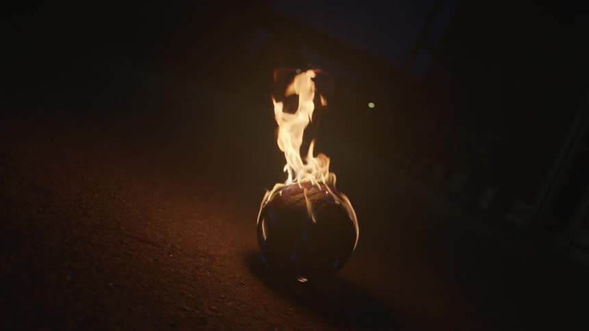 A basketball is engulfed in bright orange flames as it sits on the pavement of a dark street at night in slow motion