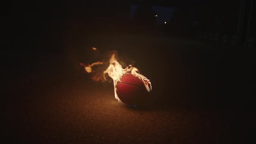 A dramatic slow-motion shot of a basketball engulfed in flames on the pavement at night