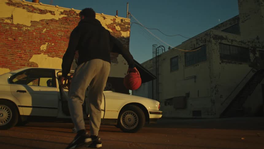 In a cinematic slow-motion shot, a young man dribbles a basketball in a gritty urban alley at sunset while a woman watches from a vintage car