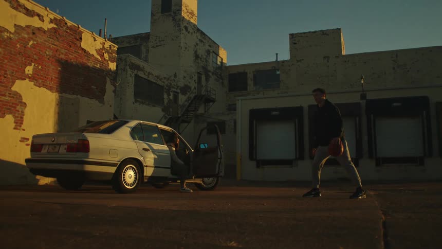 In the warm glow of sunset, a man practices dribbling a basketball in a gritty industrial parking lot as a woman watches from a nearby car