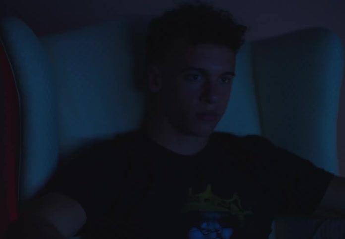 A teenage boy is captivated by a screen in a dark room, his face illuminated by the cool blue light as the camera slowly pushes in