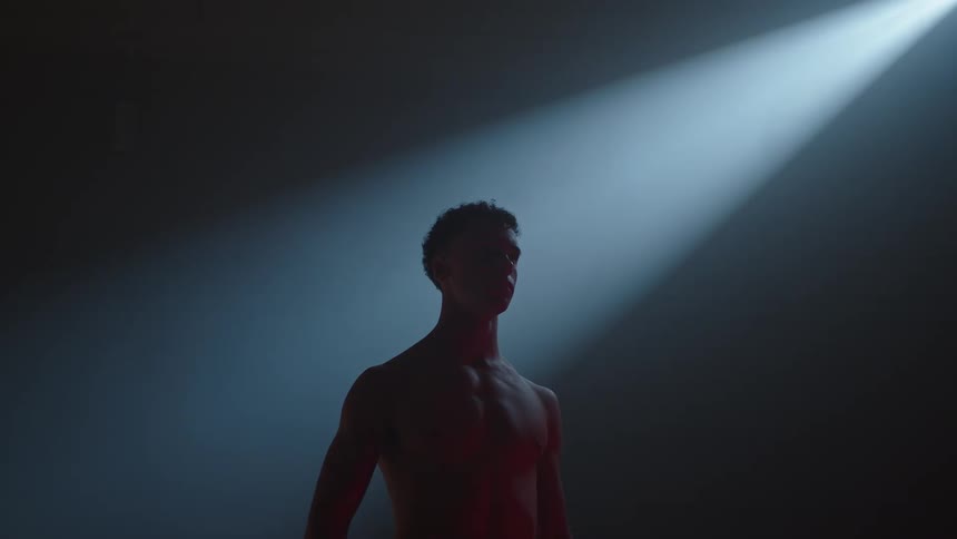 A silhouetted, muscular man stands motionless in a dramatic spotlight in a dark room