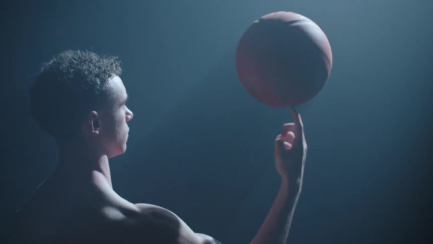 A shirtless, athletic young man skillfully spins a basketball on his finger in a dark room under a single spotlight